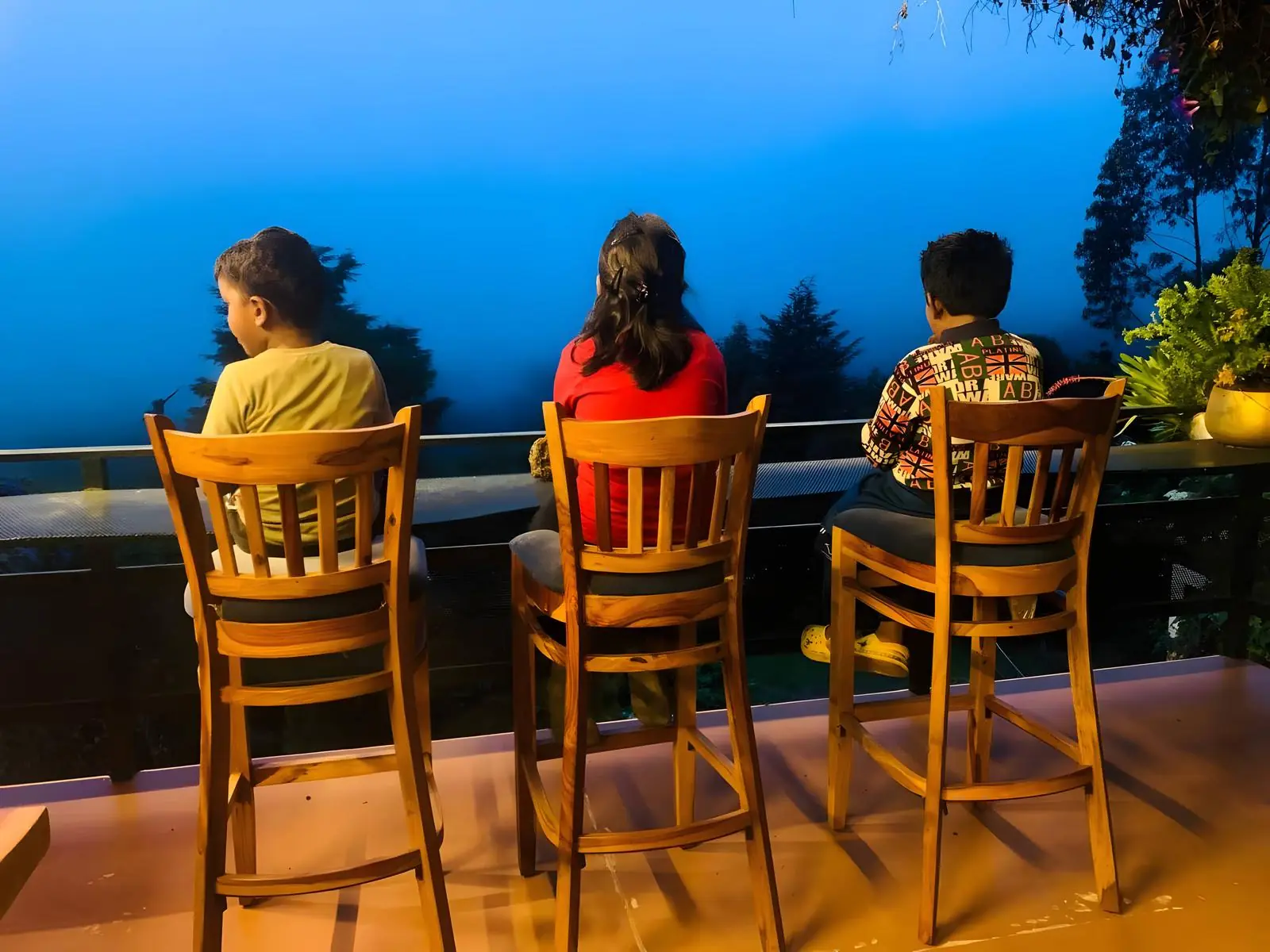 Three children on high chairs at the restaurant rail, looking out over the hills at dusk